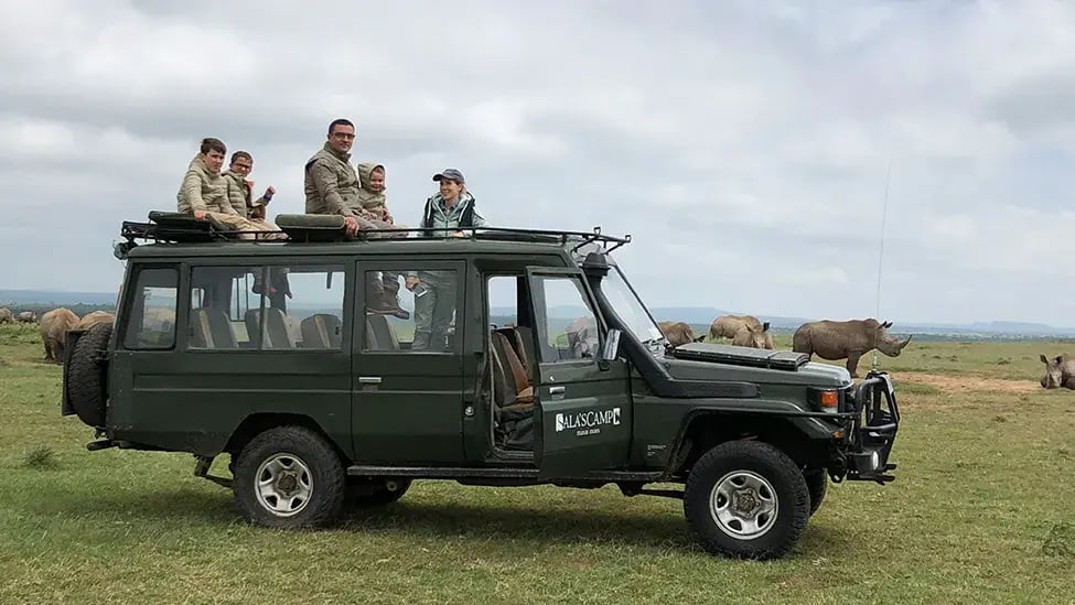 Eine Familie mit drei Söhnen sitzen auf einem Safari-Jeep. Sie sind umgeben von einer Horde Nashörnern.