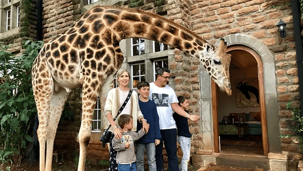 Eltern mit drei Söhnen stehen vor einem Gebäude. Neben ihnen steht ganz nah eine Giraffe und beugt den Hals über sie.