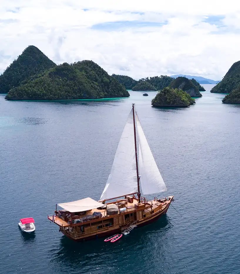 Ein Segelschiff mit weißen Segeln liegt im Meer vor einer Gruppe kleiner, grüner Inseln vor Anker