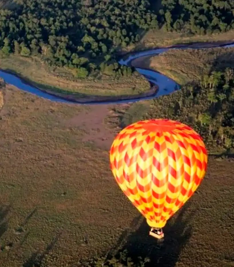 Ein rotgelber Heißluftballon ist von oben zu sehen, während er über eine idyllische Landschaft mit einem geschwungenen Fluss fährt
