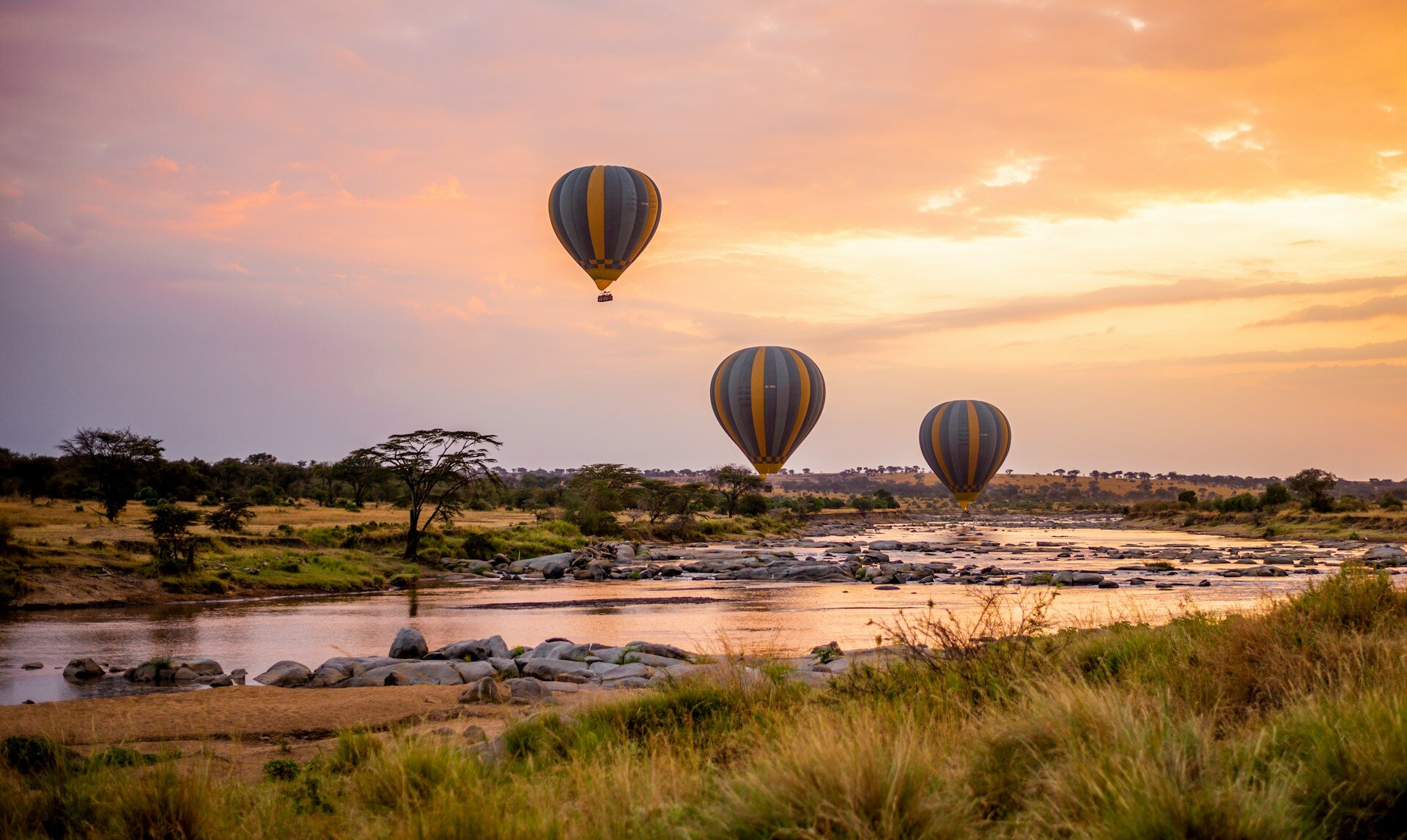 Drei Heißluftballone fahren über einen Fluss in der Serengeti bei Sonnenaufgang.