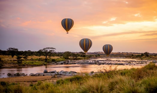 Drei Heißluftballone fahren über einen Fluss in der Serengeti bei Sonnenaufgang.