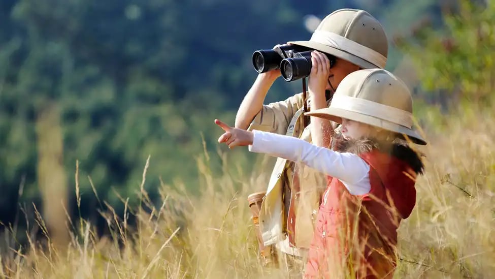 Zwei Kinder, die Safari-Hüte tragen und mit einem Fernglas in hohem Gras stehend die Umgebung beobachten
