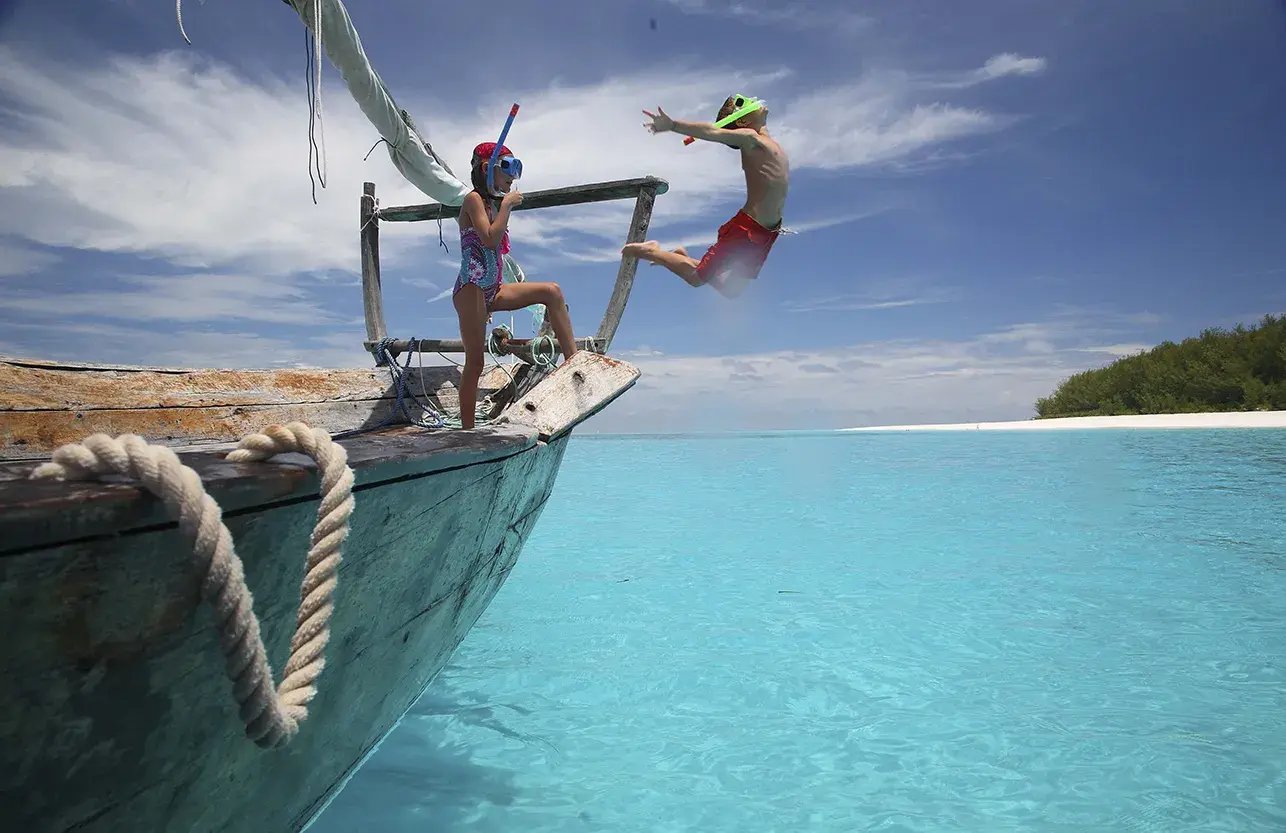 Kinder springen von einem Boot in ein türkisblaues Meer. In der Ferne ist der Strand von Mnemba Island in Sansibar zu sehen.