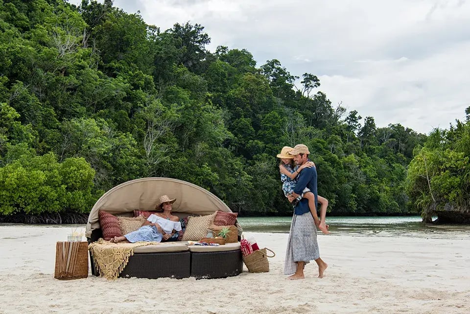 Eine Mutter mit Sonnenhut liegt auf einer gemütlichen Liege am Strand in Indonesien, während der Vater die Tochter auf dem Arm trägt und zur Mutter geht.