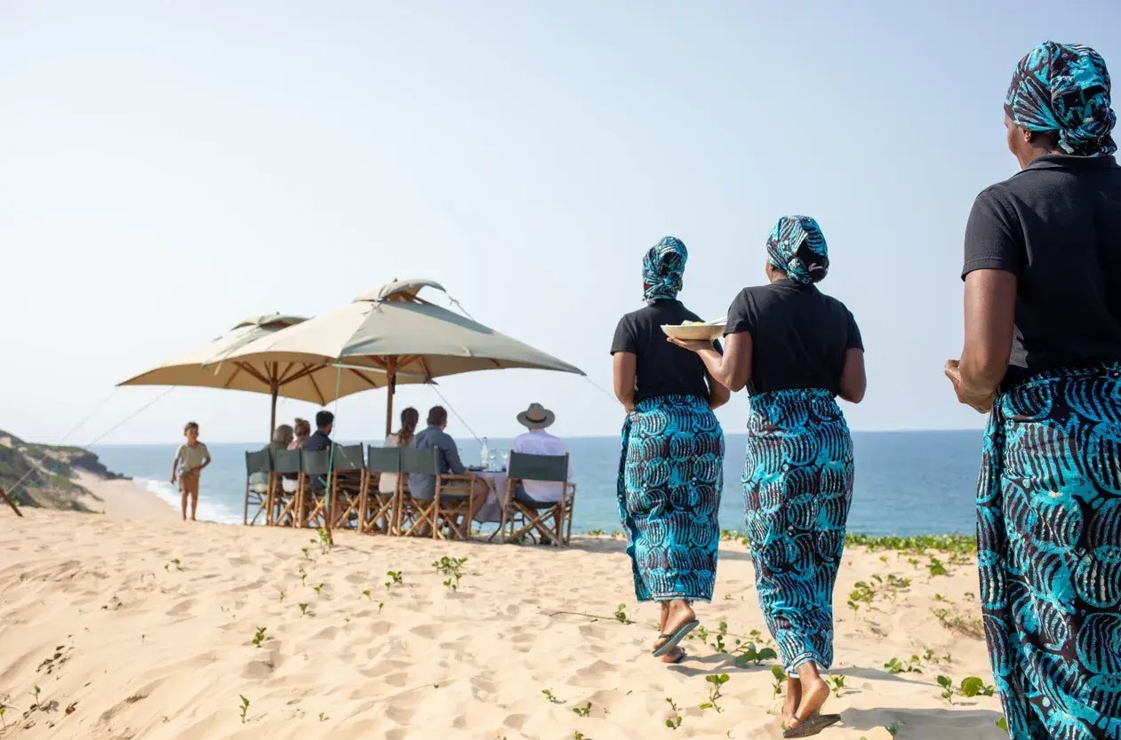 Eine Familie auf einer Mosambik-Reise sitzt unter Sonnenschirmen am Sandstrand, während das Servicepersonal das Essen bringt.