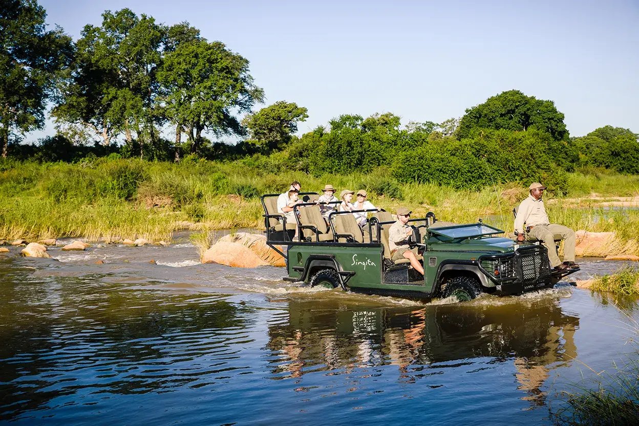 Eine Familie auf einer Luxusreise nach Singita ist auf Safari und fährt mit einem großen Jeep durch einen Fluss.