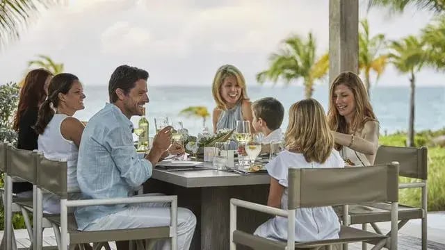 Eine Familie in einer paradiesischen Urlaubsumgebung am Meer genießt lachend das Dinner.