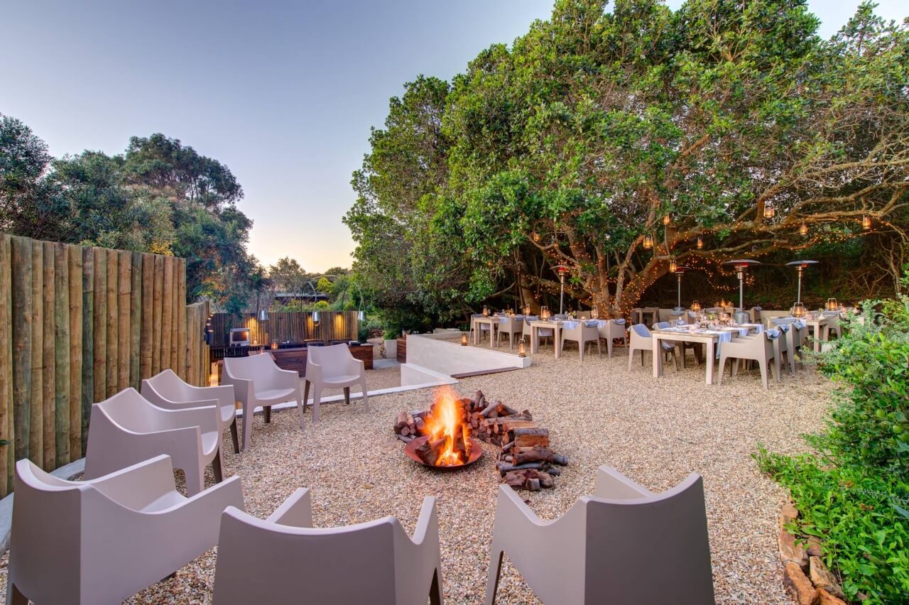 Feuerstelle mit Stühlen drumherum und einem Gartenrestaurant im Hintergrund.