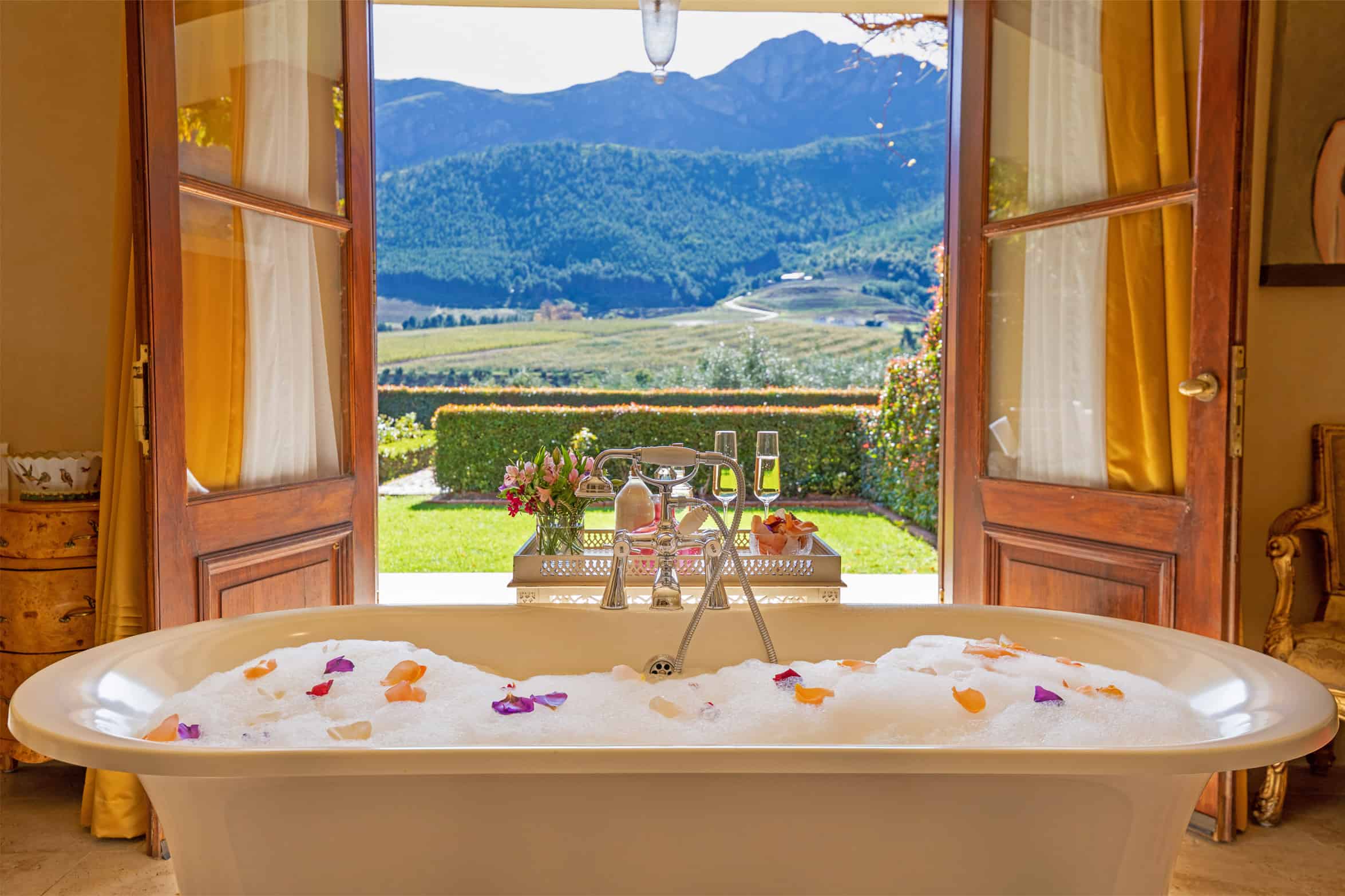 Eine Badewanne mit Schaum und Blattern steht am offenen Fenster mit Blick auf eine grüne Landschaft.