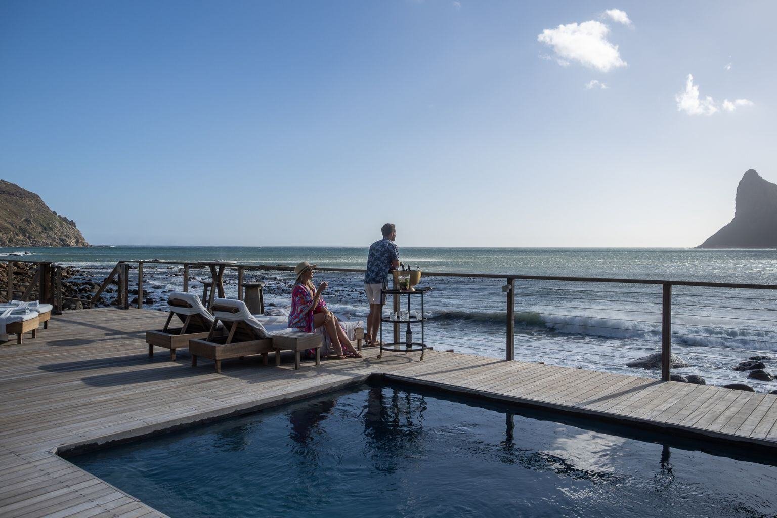 Ein Pool direkt am Meer, zwei Personen stehen an einem Geländer und genießen die Aussicht.
