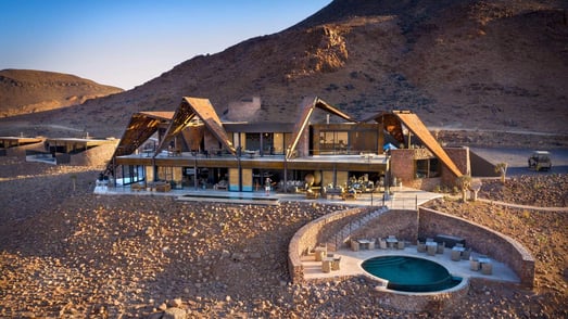 Eine Lodge mit Swimmingpool in einer rauen Felswüste vor einem Hügel.