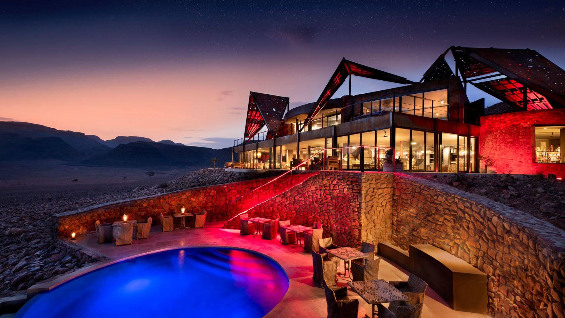 Das beleuchtete Haupthaus mit einer Lodge und dem Pool im Vordergrund.