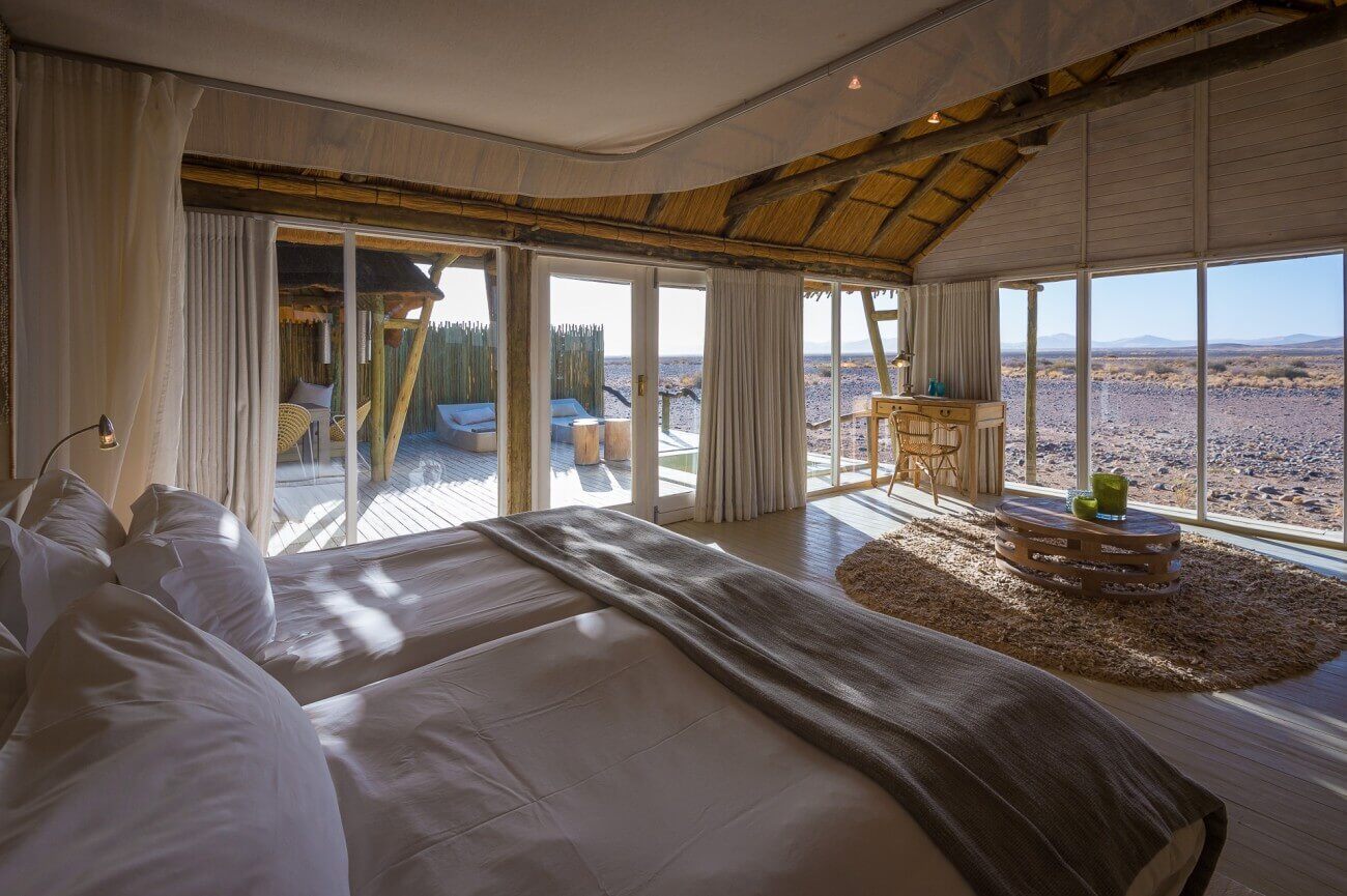 Ausblick vom großen Bett einer Lodge durch Fenster in die Wüste.