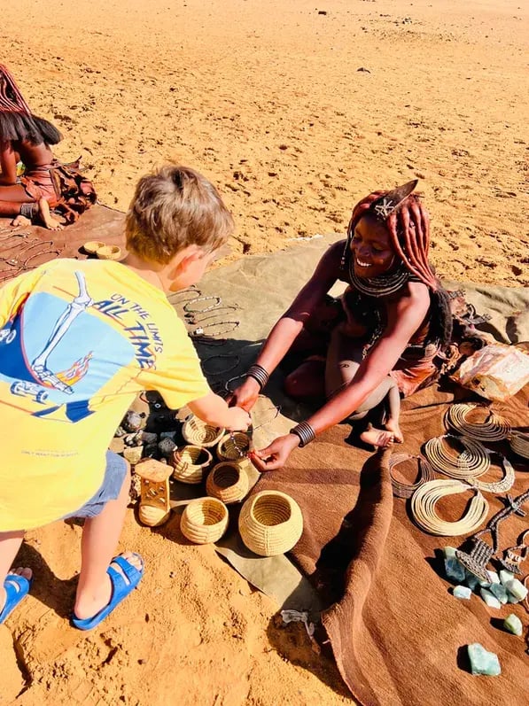 Eine Himba-Frau mit Waren bindet einem weißen Jungen ein Armband um.