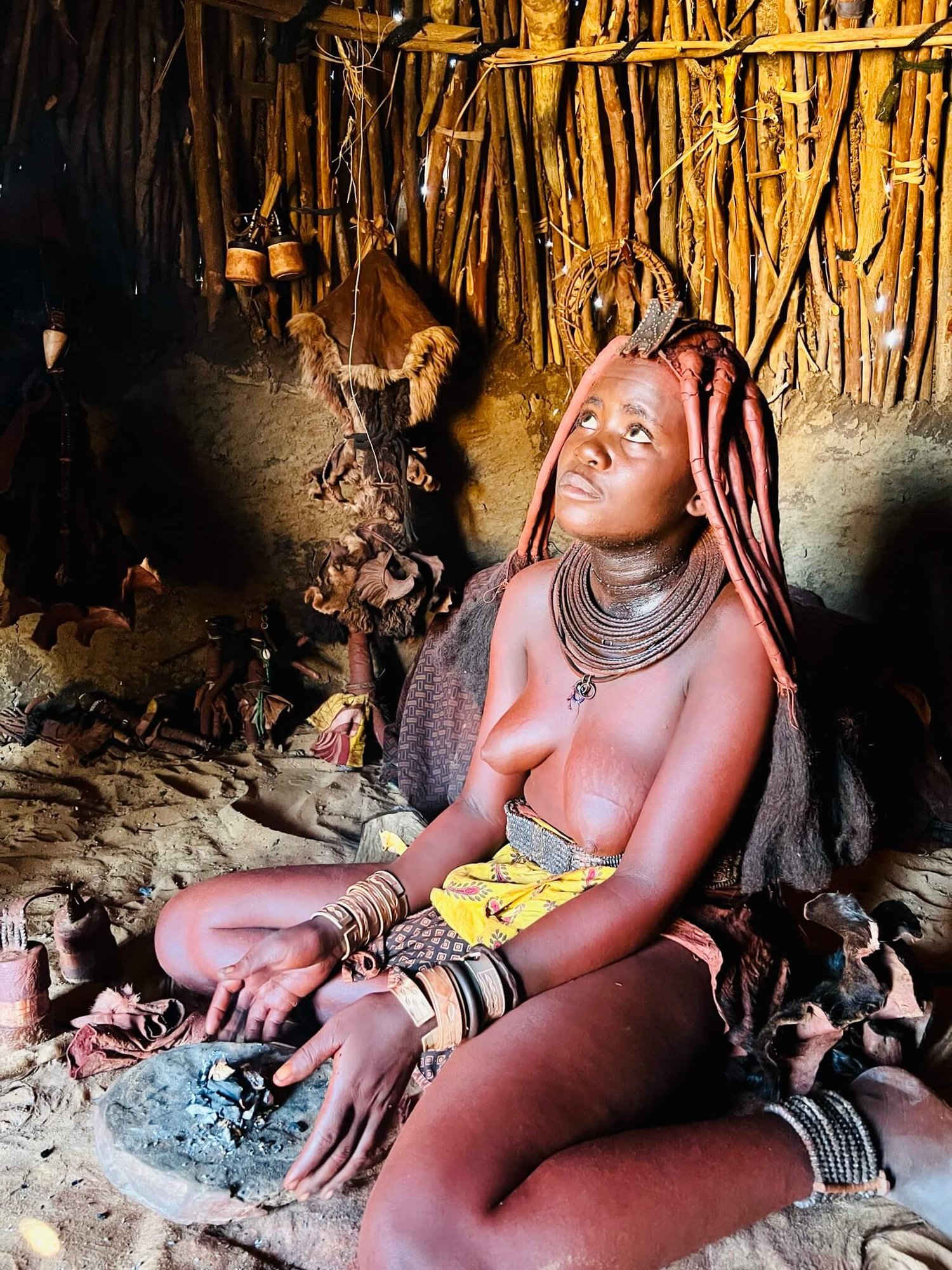 Eine Himba-Frau mit prächtiger Haarpracht sitzt auf dem Boden ihrer Lehmhütte.