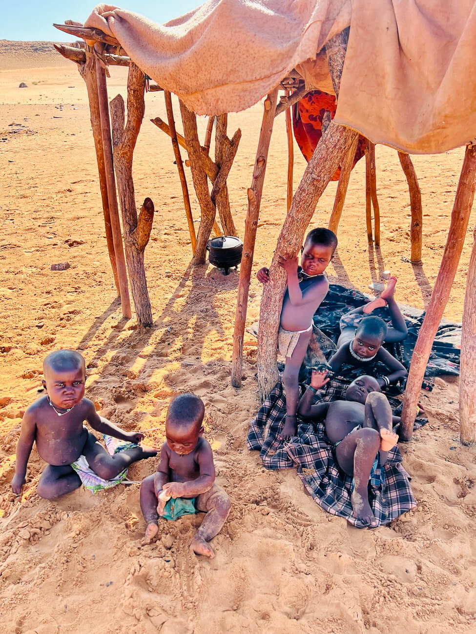 Kinder der Himba sitzen im Kaokoveld, Namibia, unter einem Sonnenschutz aus natürlichen Materialien.