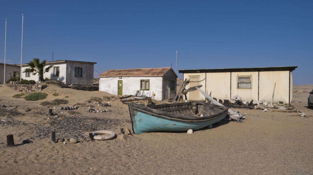 Ein türkises Fischerboot liegt am Strand vor drei Hütten.