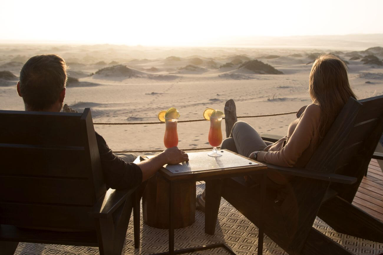 Ein Paar sitzt auf einer Terrasse mit Cocktails und blickt auf die vor ihnen liegende Wüstenlandschaft.