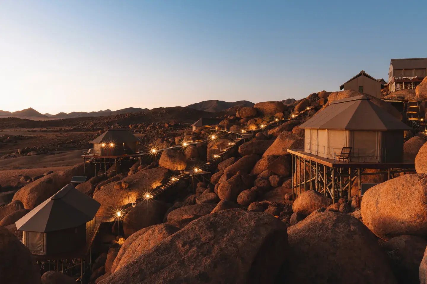 Die Unterkünfte der Sonop Lodge bei Sonnenuntergang am Felsen, die ersten Beleuchtungen sind an.
