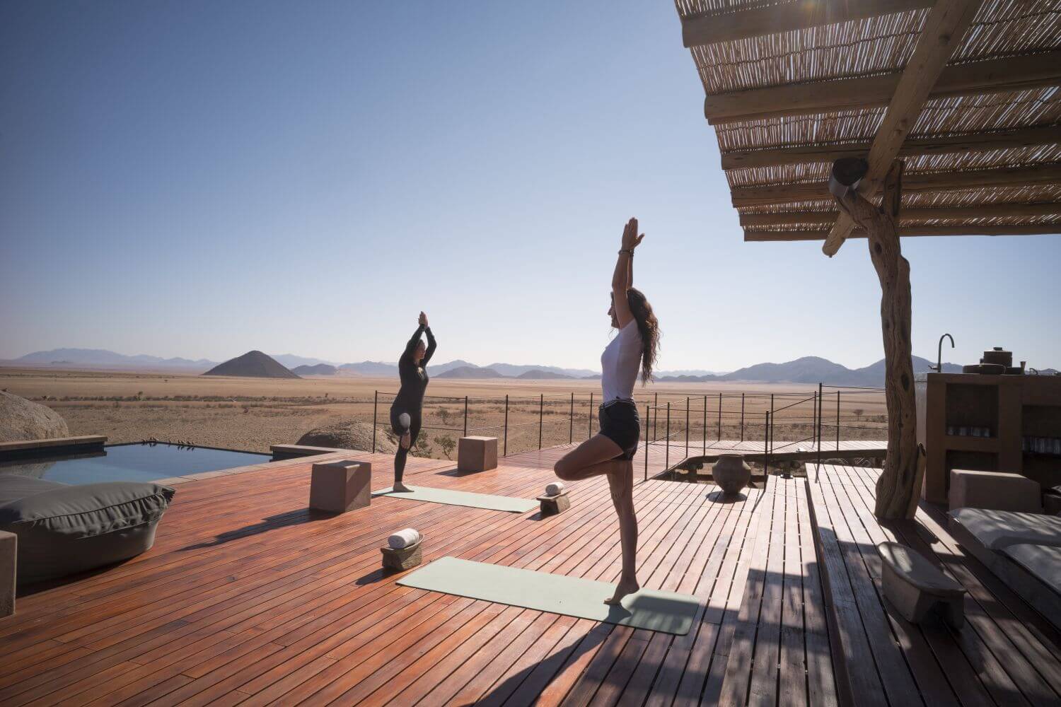 Zwei Frauen machen Yoga auf einer Holzveranda mit Blick in die Wüste.