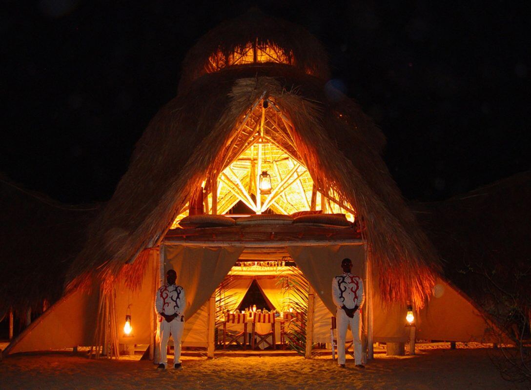 Hell erleuchtete Lodge am Abend, davor stehen zwei Mitarbeiter.