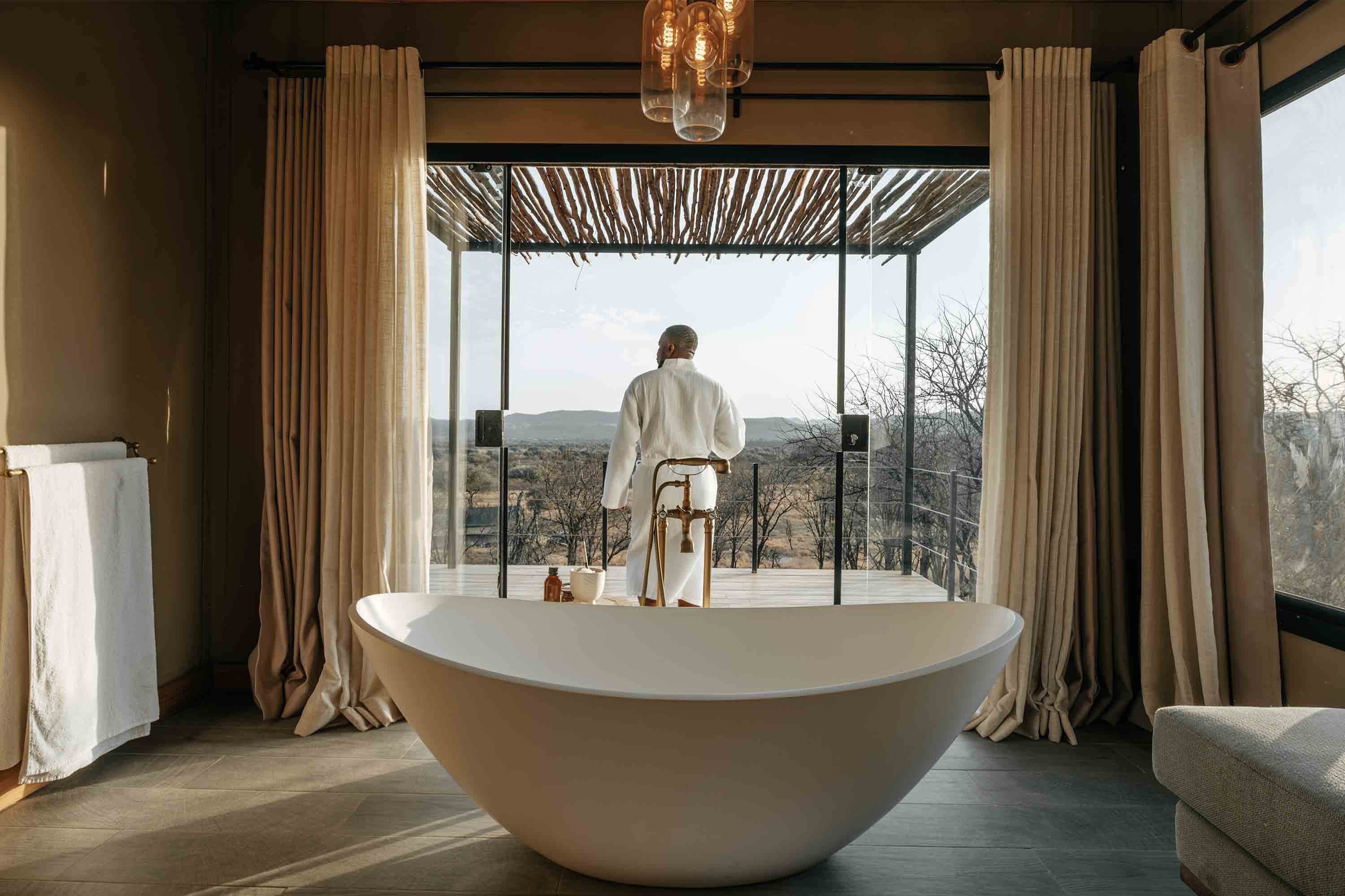 Ein Mann im Bademantel steht auf einem Balkon, im Vordergrund ist eine freistehende Badewanne.
