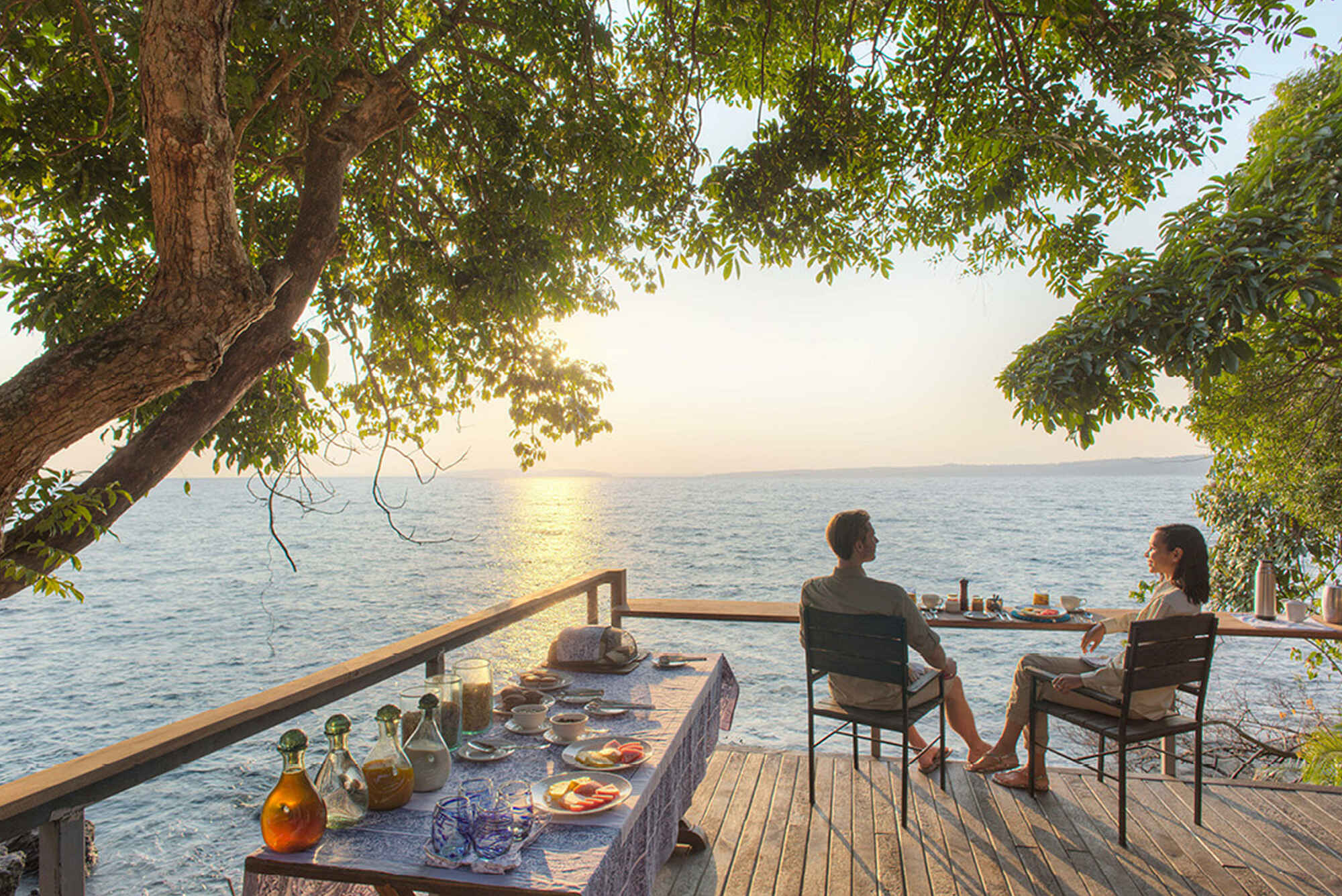 Ein Paar sitzt bei Sonnenuntergang auf einer Terrasse am See, hinter ihnen steht ein Tisch mit Getränken und Snacks.