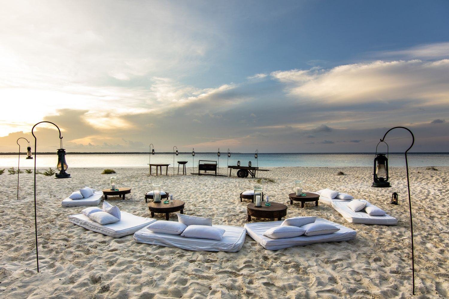 Weiße Kissen liegen am Strand, Laternen stehen im Sand - alles ist für einen Sonnenuntergang vorbereitet.