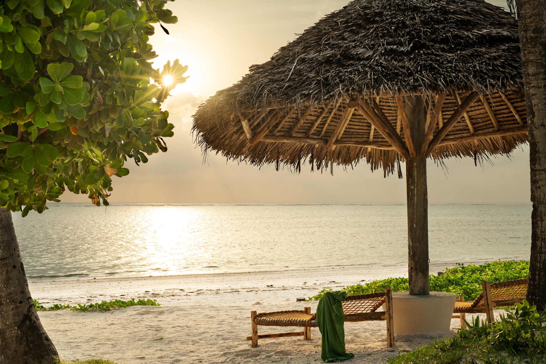 Tiefstehende Sonne am Strand, an dem zwei Liegen und ein Sonnenschirm aus Stroh stehen.