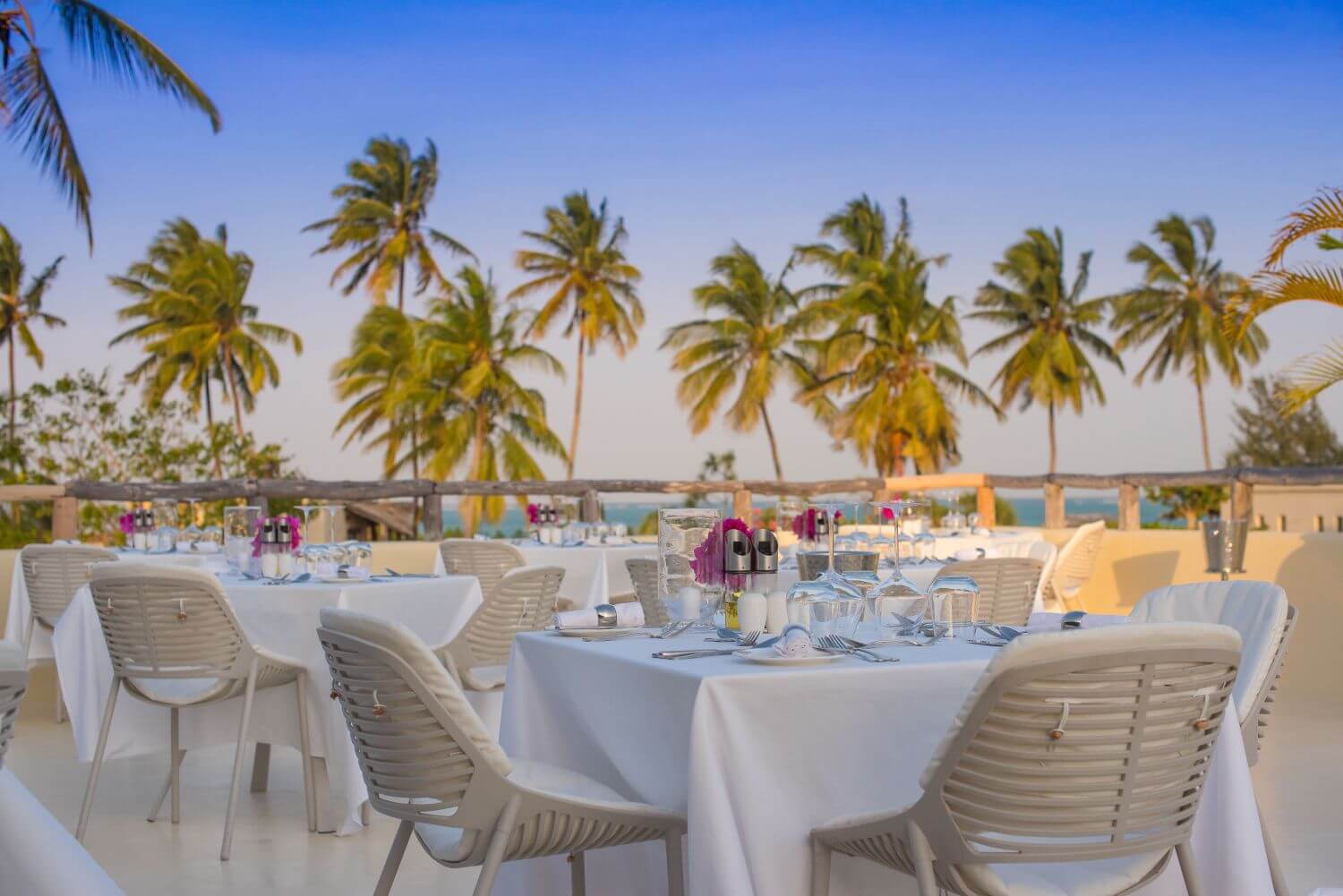 Edles Restaurant im Freien, Tische und Stühle sind in Weiß gehalten, im Hintergrund wiegen sich Palmen im Wind.