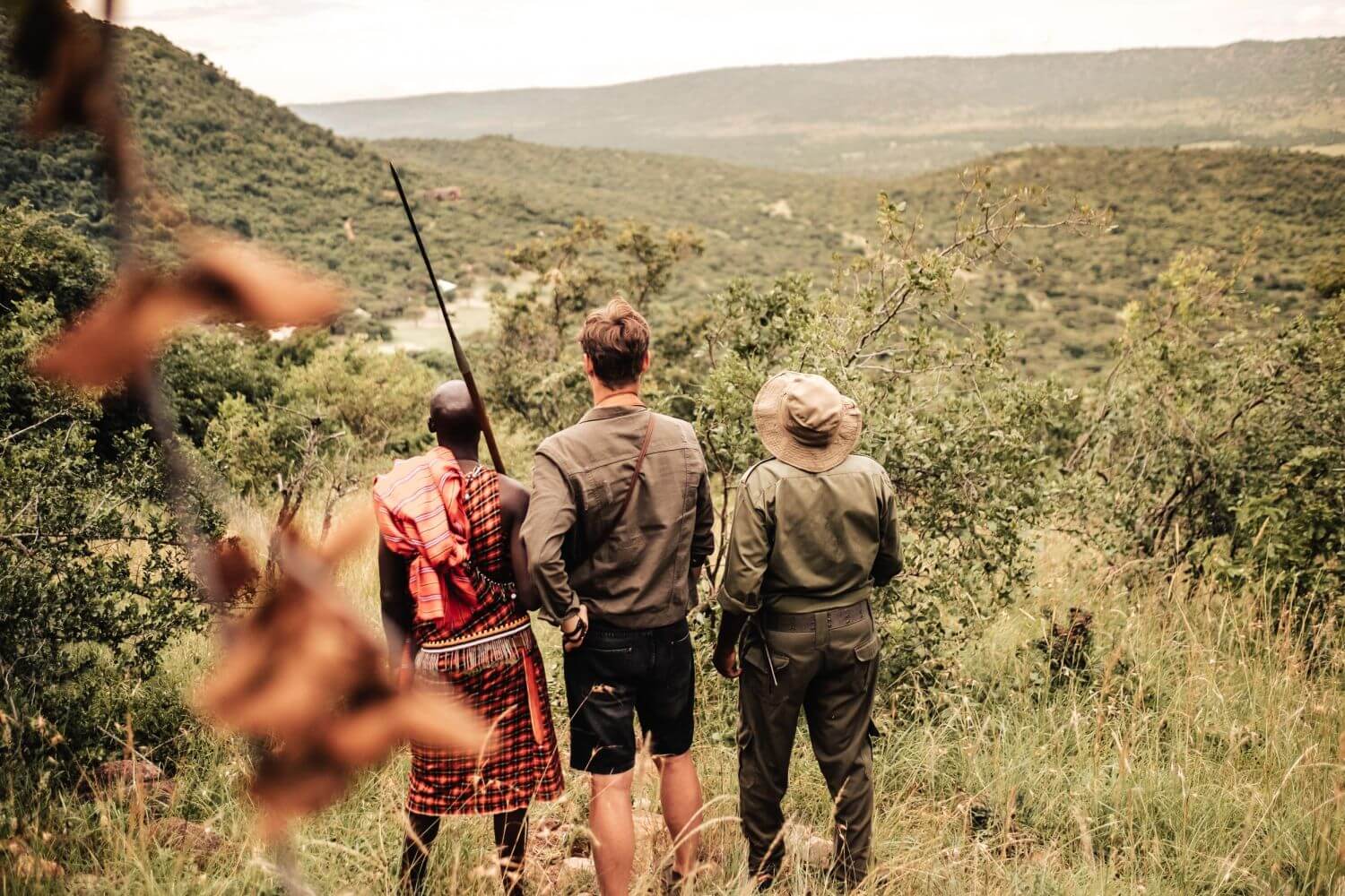 Ein Maasai und zwei Gäste blicken bei einem Naturewalk herunter auf einen Fluss.