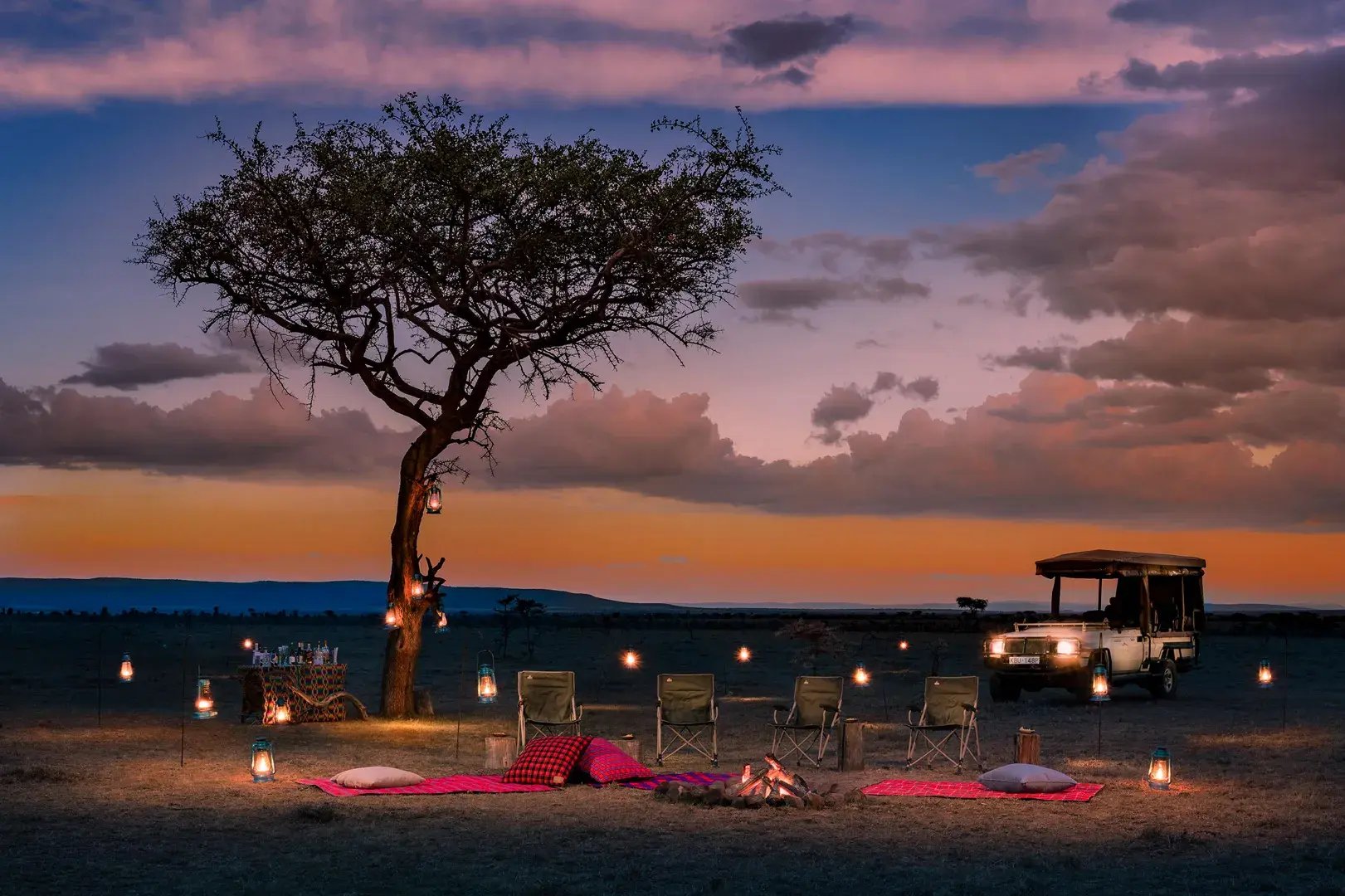 Sundowner unter einem Baum. Um eine Picknickdecke herum sind Lichter aufgestellt.