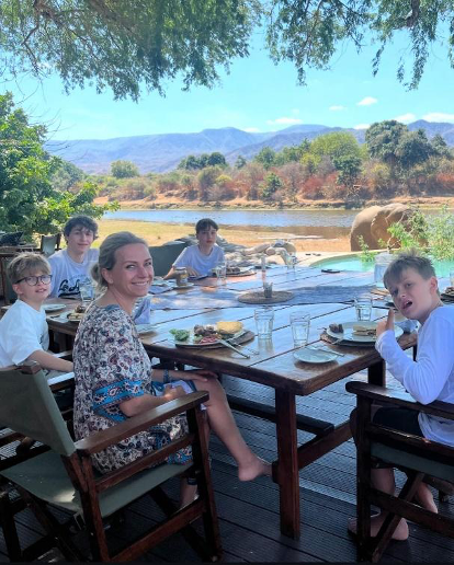 Eine blonde Frau sitzt mit vier Jungen beim Essen an einem großen Tisch aus Holz im Chongwe House. Alle fünf Personen schauen in die Kamera. Im Hintergrund ist eine afrikanische Landschaft mit einem Fluss und einem Elefanten zu sehen.