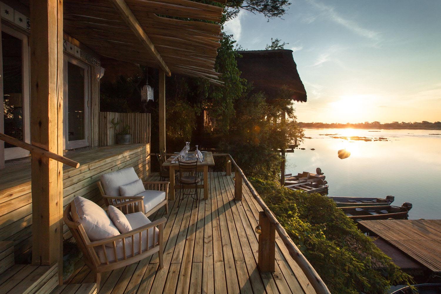 Terrasse mit gepolsterten Stühlen. Die Sonne geht über einem Fluss unter.