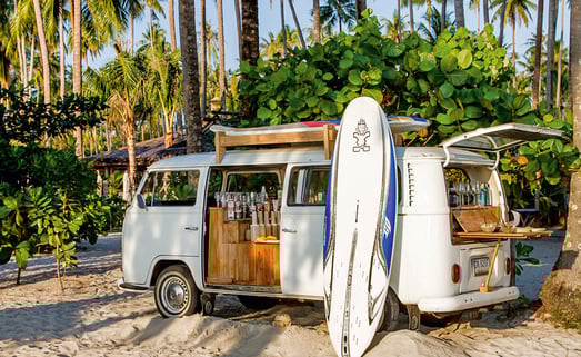 Ein nostalgischer VW-Bus, der im Inneren zu einer Cocktailbar umgebaut wurde, steht am Sandstrand unter Palmen