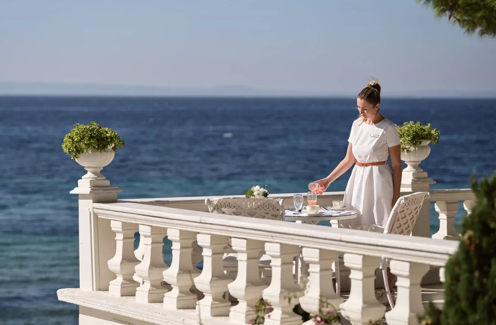 Eine Frau steht an einem Tisch an der Ecke einer Terrasse mit unendlichem Blick auf das Meer.