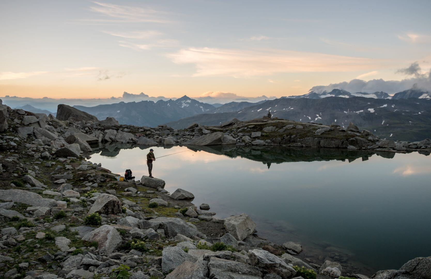 Ein Angler steht bei Morgengrauen an einem Bergsee.