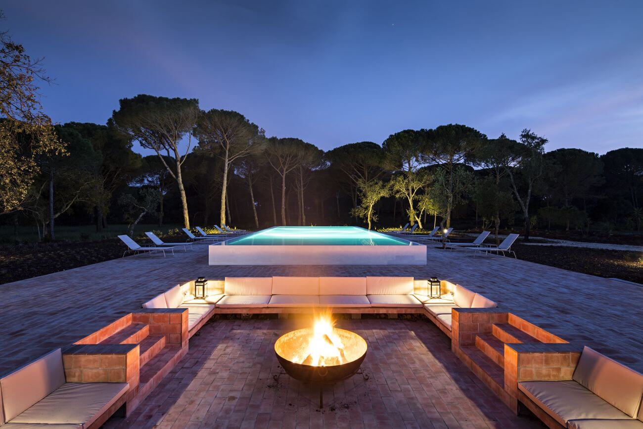 Feuerschale am Abend mit beleuchtetem Pool im Hintergrund.