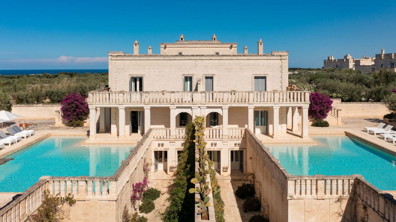 Weißes Hauptgebäude eines apulischen Anwesens, zu deren rechter und linker Seite jeweils ein Pool ist.