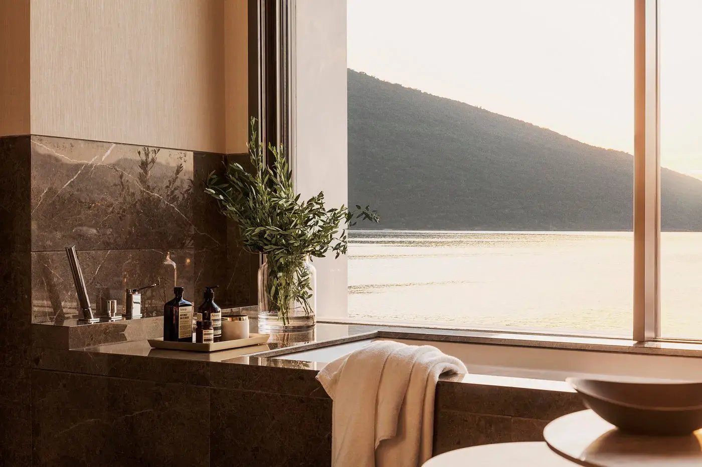 Wellnessprodukte im Hotelzimmer und ein Handtuch. Von einer Badewanne aus blickt man auf einen See.