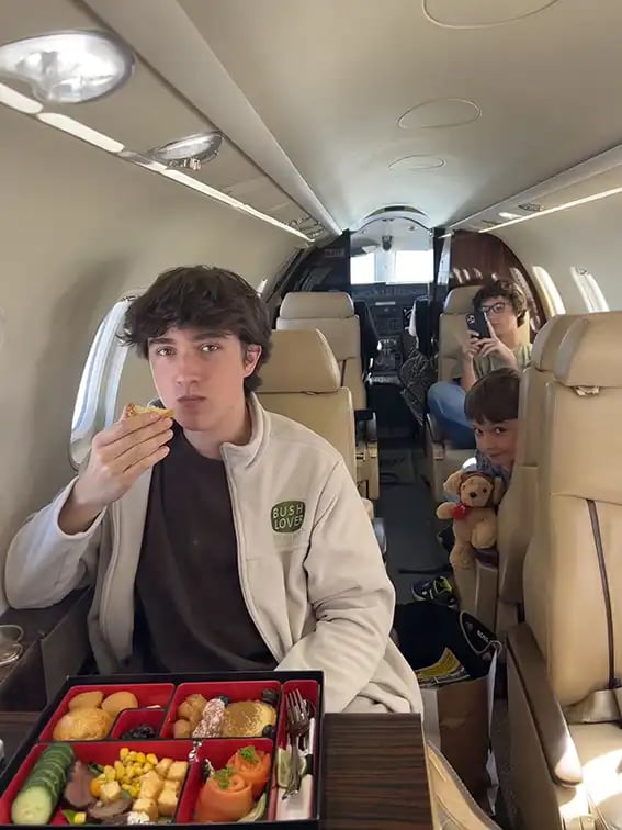 Zwei Jungs und ein Teenager fliegen in einem Privatjet. Der Teenager hat vor sich eine Platte mit gesundem, leckeren Essen.