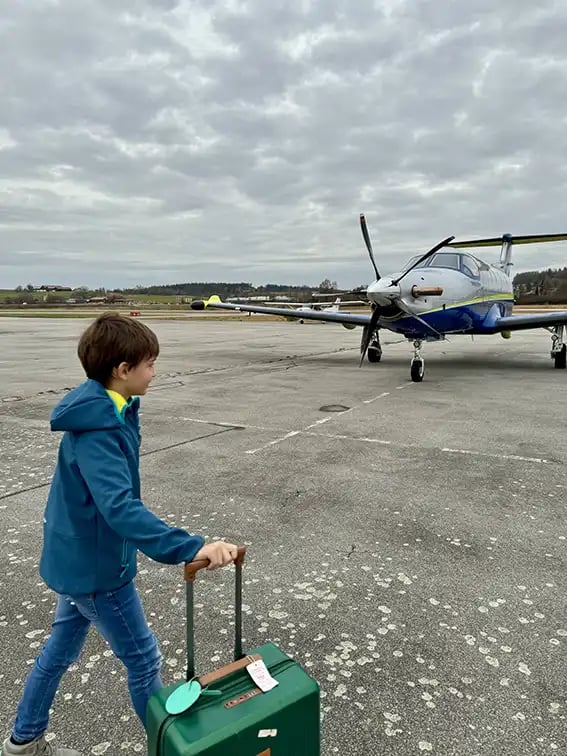 Ein kleiner Junge läuft mit seinem Rollkoffer über eine Landebahn zu einem Privatjet.
