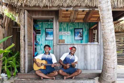 Zwei Einheimische sitzen vor einer Hütte aus Holz neben einer Palme und machen auf einer Gitarre und einer Ukulele Musik